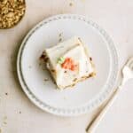 A slice of frosted cake with decorative icing sits on a white plate next to a fork; a small bowl of chopped nuts is nearby.