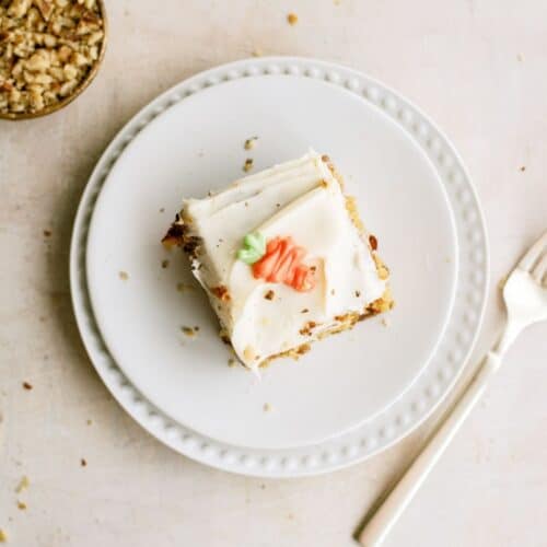 A slice of frosted cake with decorative icing sits on a white plate next to a fork; a small bowl of chopped nuts is nearby.