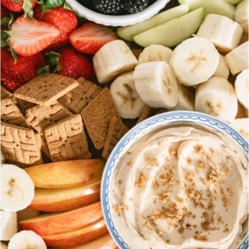 A platter with sliced bananas, apples, strawberries, cucumbers, graham crackers, blackberries, and a bowl of creamy dip topped with brown sugar.