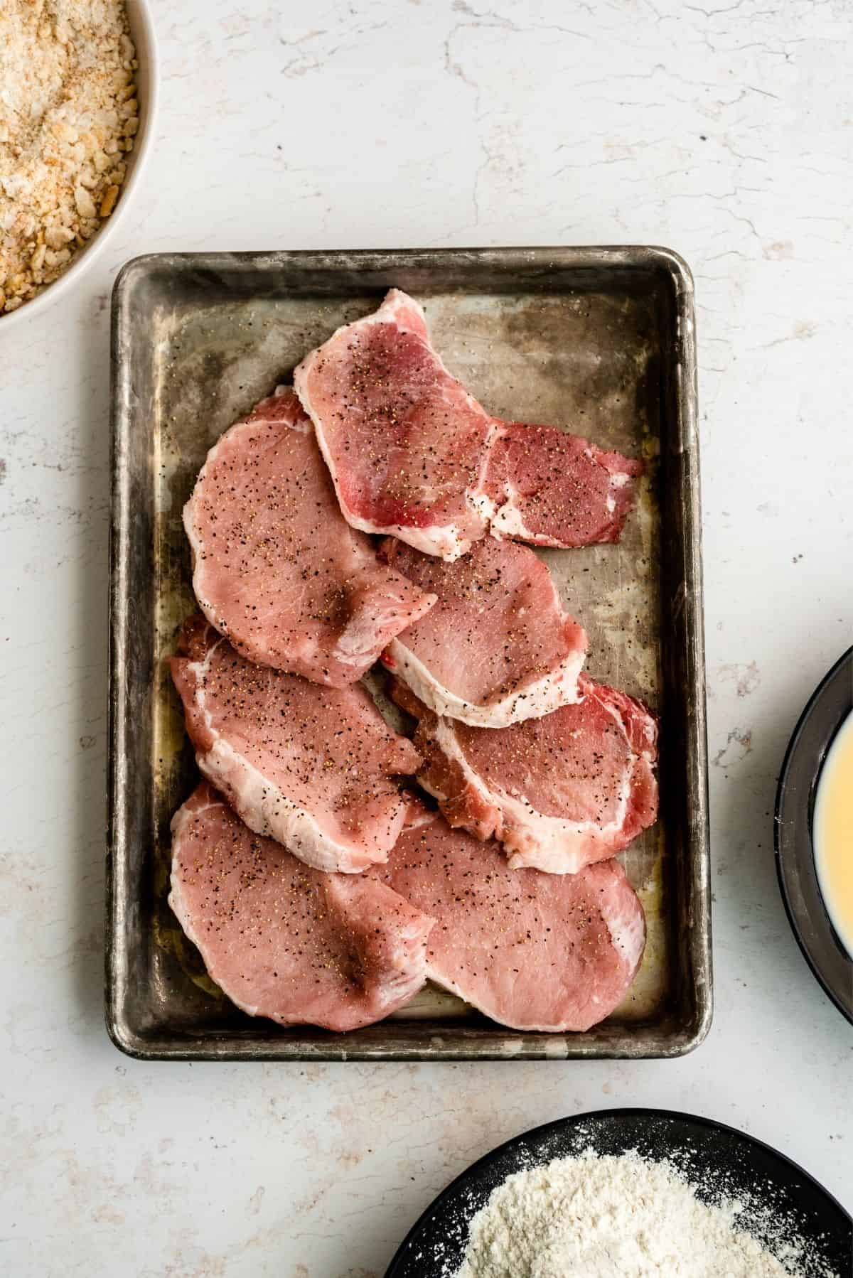 Raw pork chops on a sheet pan with salt and pepper on them