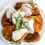A plate of breaded and fried chicken cutlets topped with white gravy and garnished with sprigs of fresh rosemary.