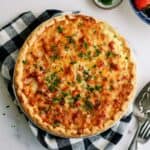 A golden-brown quiche garnished with chopped herbs sits on a checkered cloth. A pie server is beside it, with small bowls of chopped herbs and strawberries nearby.