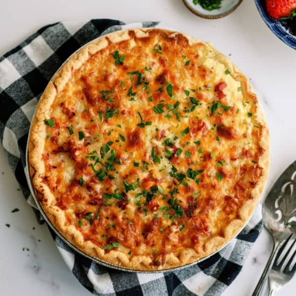A golden-brown quiche garnished with chopped herbs sits on a checkered cloth. A pie server is beside it, with small bowls of chopped herbs and strawberries nearby.