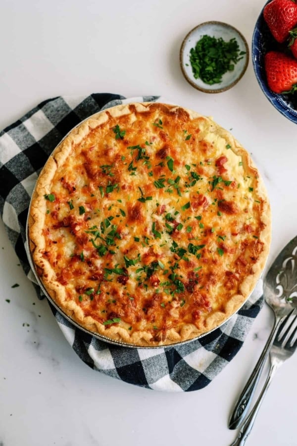 A golden-brown quiche garnished with chopped herbs sits on a checkered cloth. A pie server is beside it, with small bowls of chopped herbs and strawberries nearby.