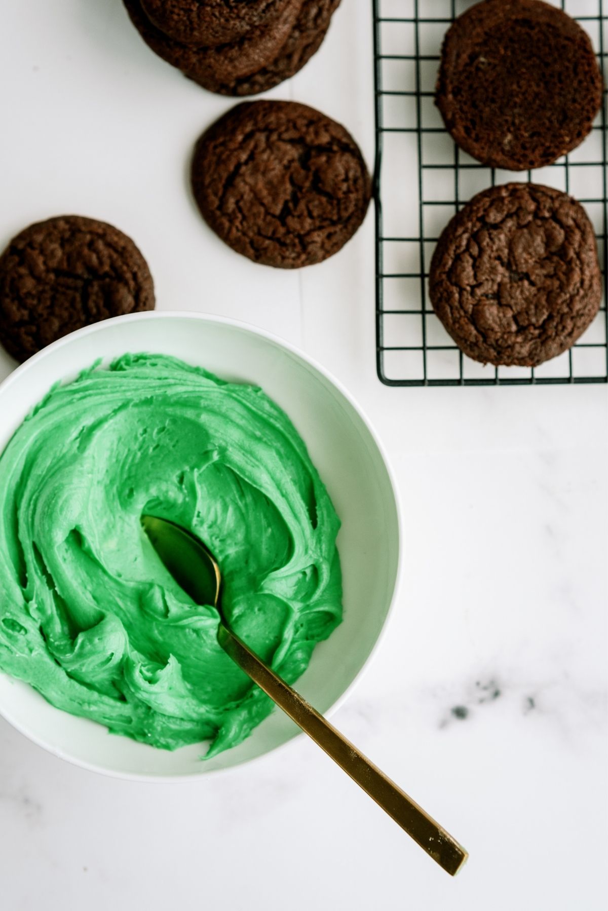 Chocolate cookies on a cooling rack with a bowl of green frosting