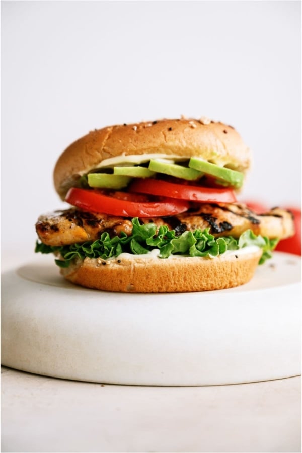Marinated Grilled Chicken Sandwich topped with tomatoes and avocados on a white background.
