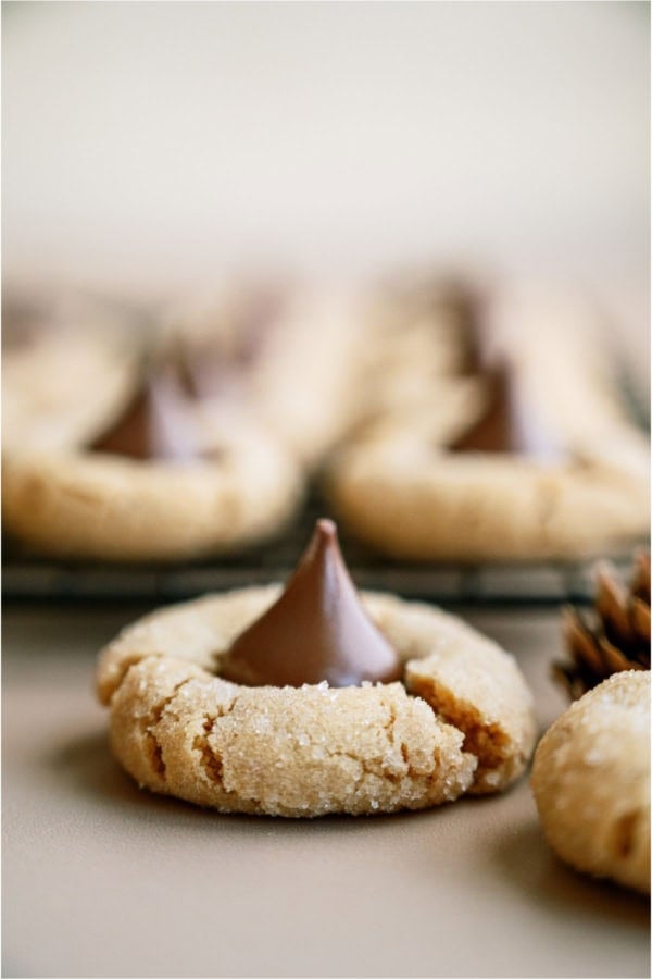 One Peanut Butter Blossom (Hershey’s Kiss Cookie) in front with remaining Peanut Butter Blossoms (Hershey’s Kiss Cookies) on a cooling rack in the background.
