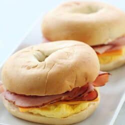 Two bagels filled with slices of ham, cheese, and scrambled eggs are placed on a white rectangular plate.