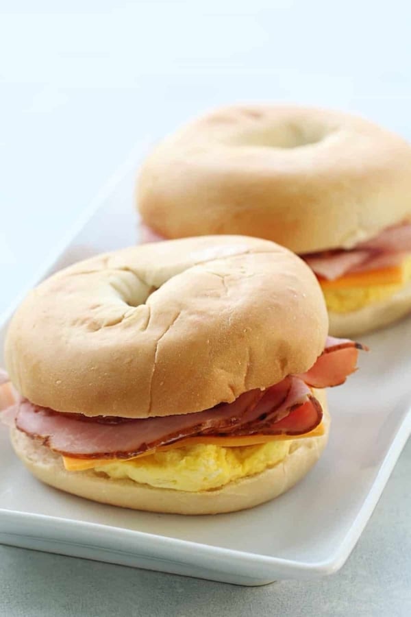 Two bagels filled with slices of ham, cheese, and scrambled eggs are placed on a white rectangular plate.