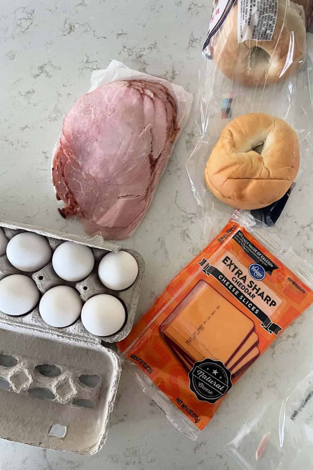 Ham slices, extra sharp cheddar cheese, six eggs in a carton, a bagel, and an open bag with another bagel on a light-colored countertop.