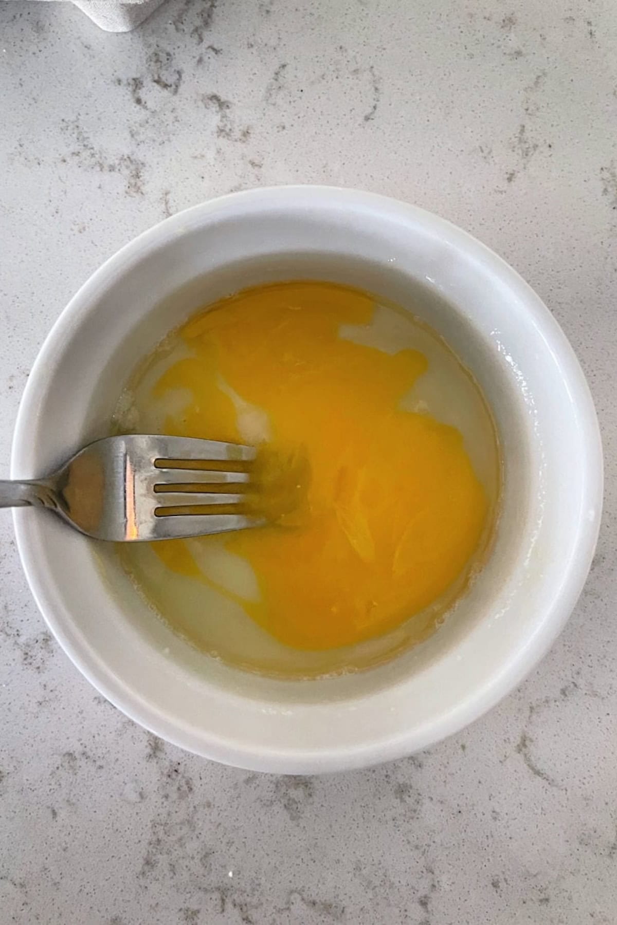 A fork rests in a white bowl containing an egg with yolk and egg white, placed on a marbled countertop.