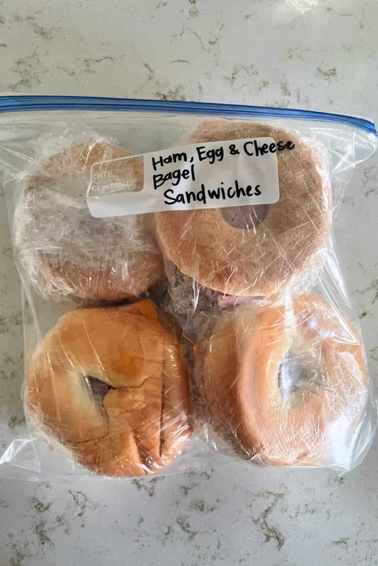 Four bagel sandwiches labeled "Ham, Egg & Cheese Bagel Sandwiches" are individually wrapped in plastic and stored in a clear resealable bag on a countertop.