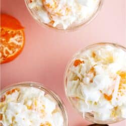 Three glass bowls filled with a creamy fruit salad containing orange segments, pineapple pieces, shredded coconut, and mini marshmallows, placed on a pink surface with spoons.