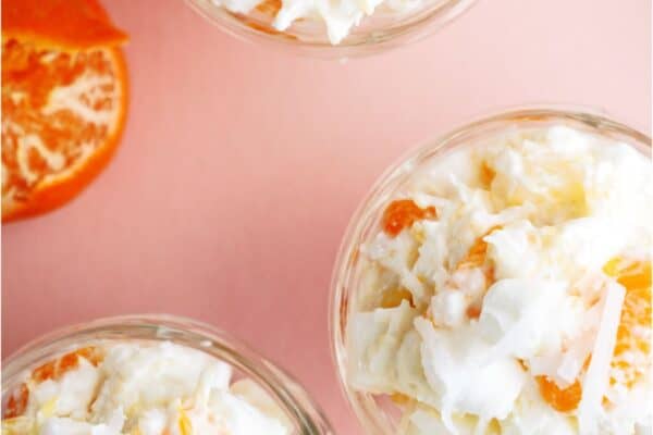 Three glass bowls filled with a creamy fruit salad containing orange segments, pineapple pieces, shredded coconut, and mini marshmallows, placed on a pink surface with spoons.
