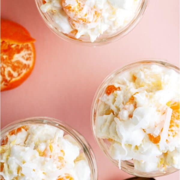 Three glass bowls filled with a creamy fruit salad containing orange segments, pineapple pieces, shredded coconut, and mini marshmallows, placed on a pink surface with spoons.