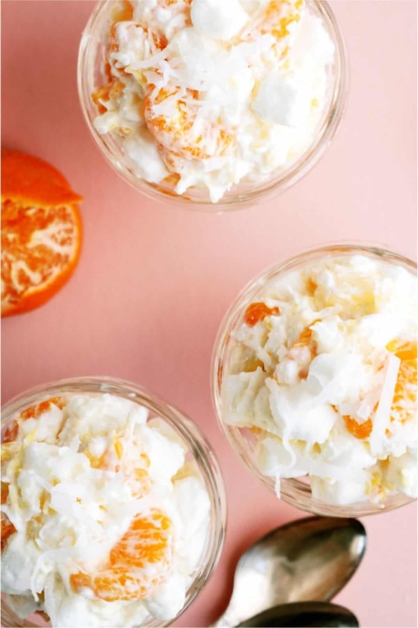 Three glass bowls filled with a creamy fruit salad containing orange segments, pineapple pieces, shredded coconut, and mini marshmallows, placed on a pink surface with spoons.