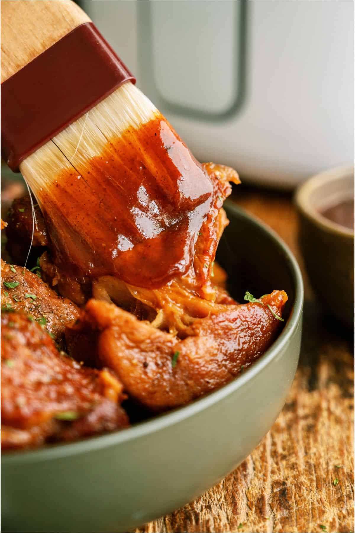 Basting brush brushing on BBQ sauce to cooked country ribs in a bowl.