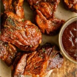 Several pieces of cooked, sauced pork country-style ribs are arranged on brown parchment paper beside a small bowl of barbecue sauce.