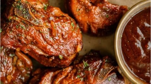 Several pieces of cooked, sauced pork country-style ribs are arranged on brown parchment paper beside a small bowl of barbecue sauce.