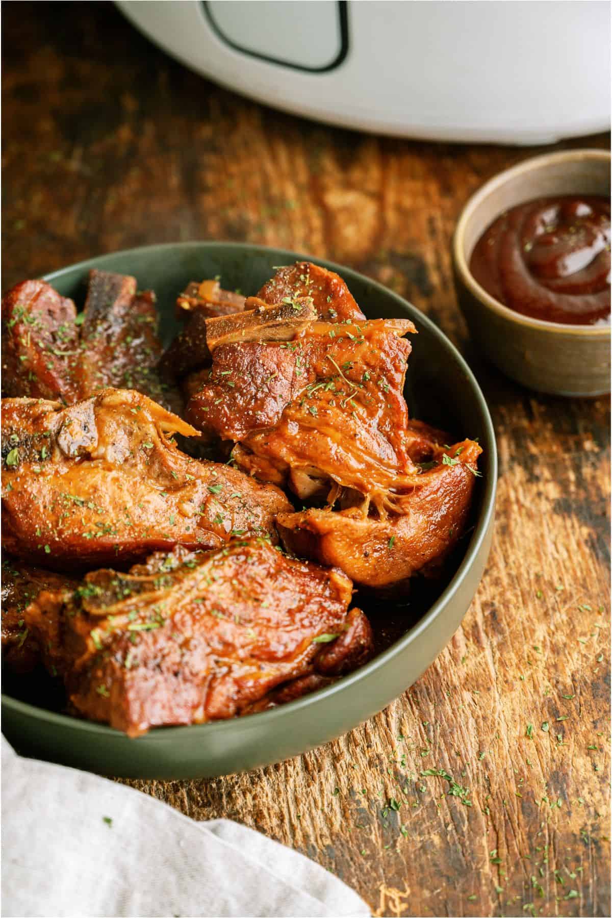 BBQ Slow Cooker Country Style Ribs in a bowl with the slow cooker in the background and a small bowl of BBQ sauce on the side.