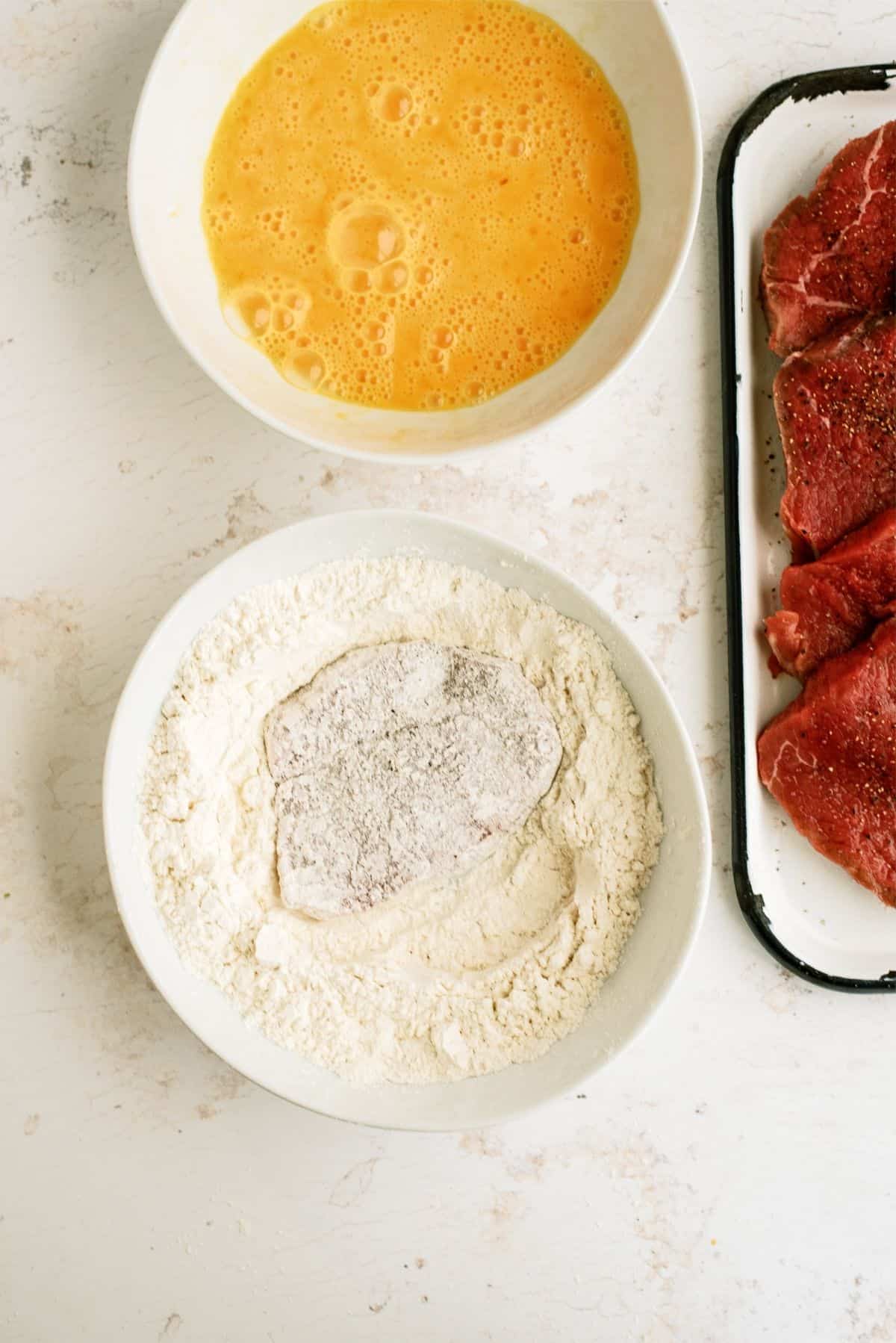 Steak with a bowl of flower and a bowl of eggs for dipping