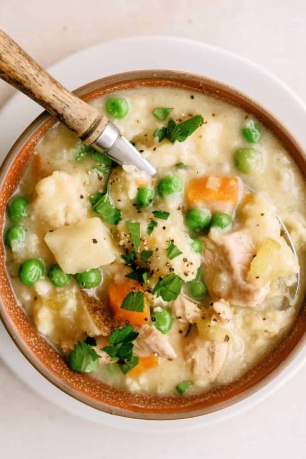 A bowl of creamy chicken stew with peas, carrots, potatoes, and herbs, served with a spoon on a white plate.