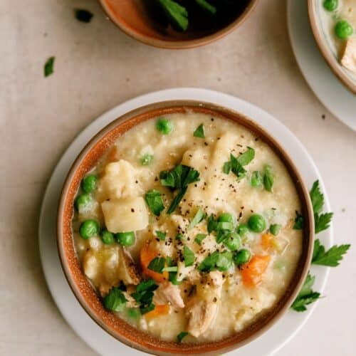 A bowl of creamy chicken and vegetable soup garnished with fresh parsley, featuring peas, carrots, potatoes, and chunks of chicken.