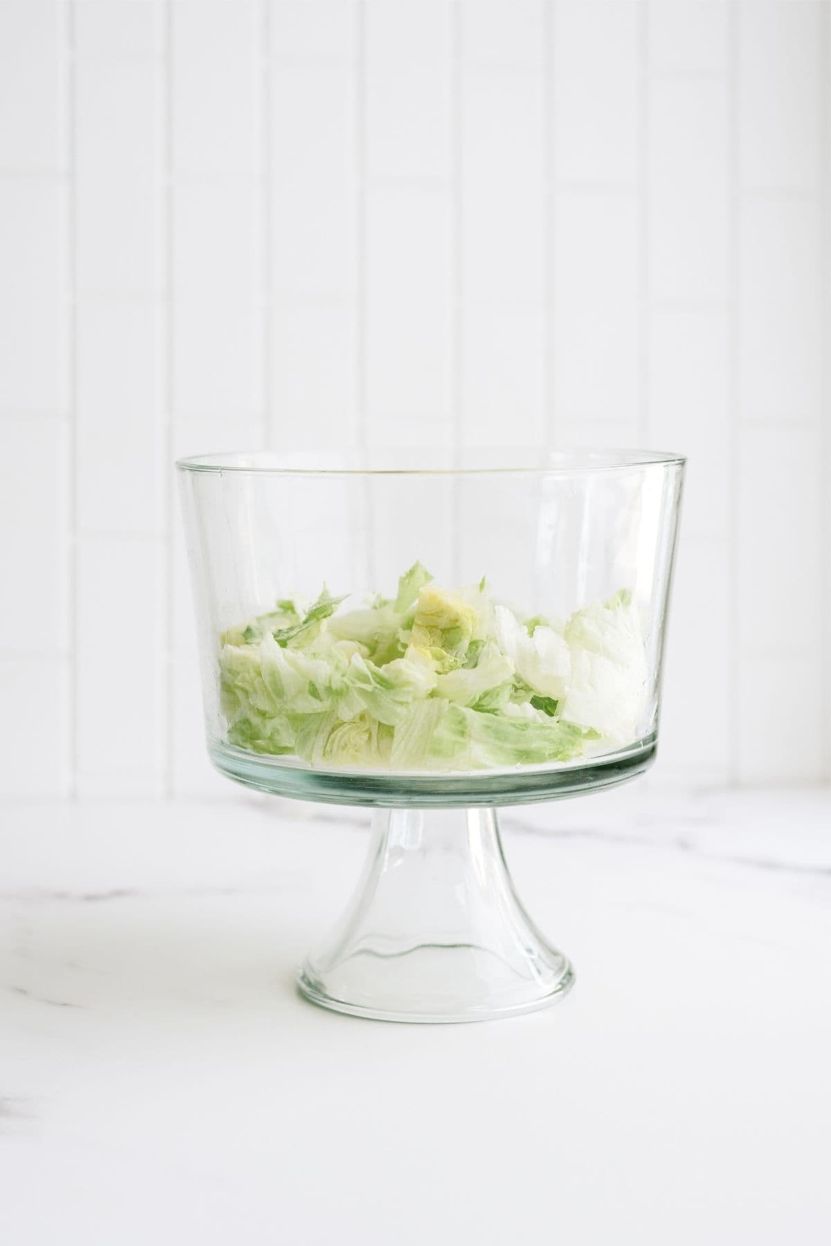 A clear glass bowl containing chopped iceberg lettuce sits on a white surface with a tiled white background.