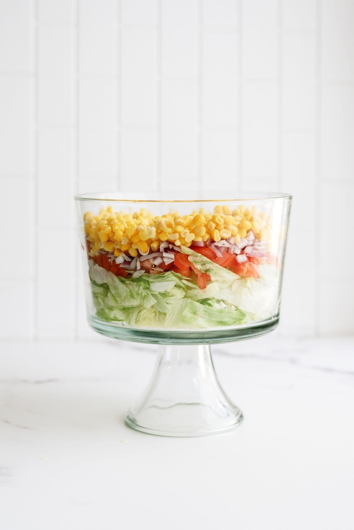 A glass trifle bowl containing a layered salad with lettuce, tomatoes, red onions, and corn.