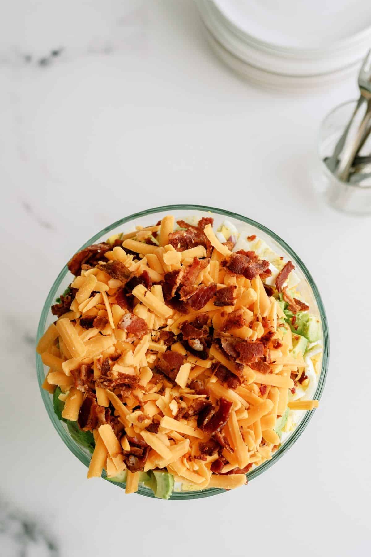 A glass bowl filled with a layered salad topped with shredded cheddar cheese and crumbled bacon, set on a white surface.