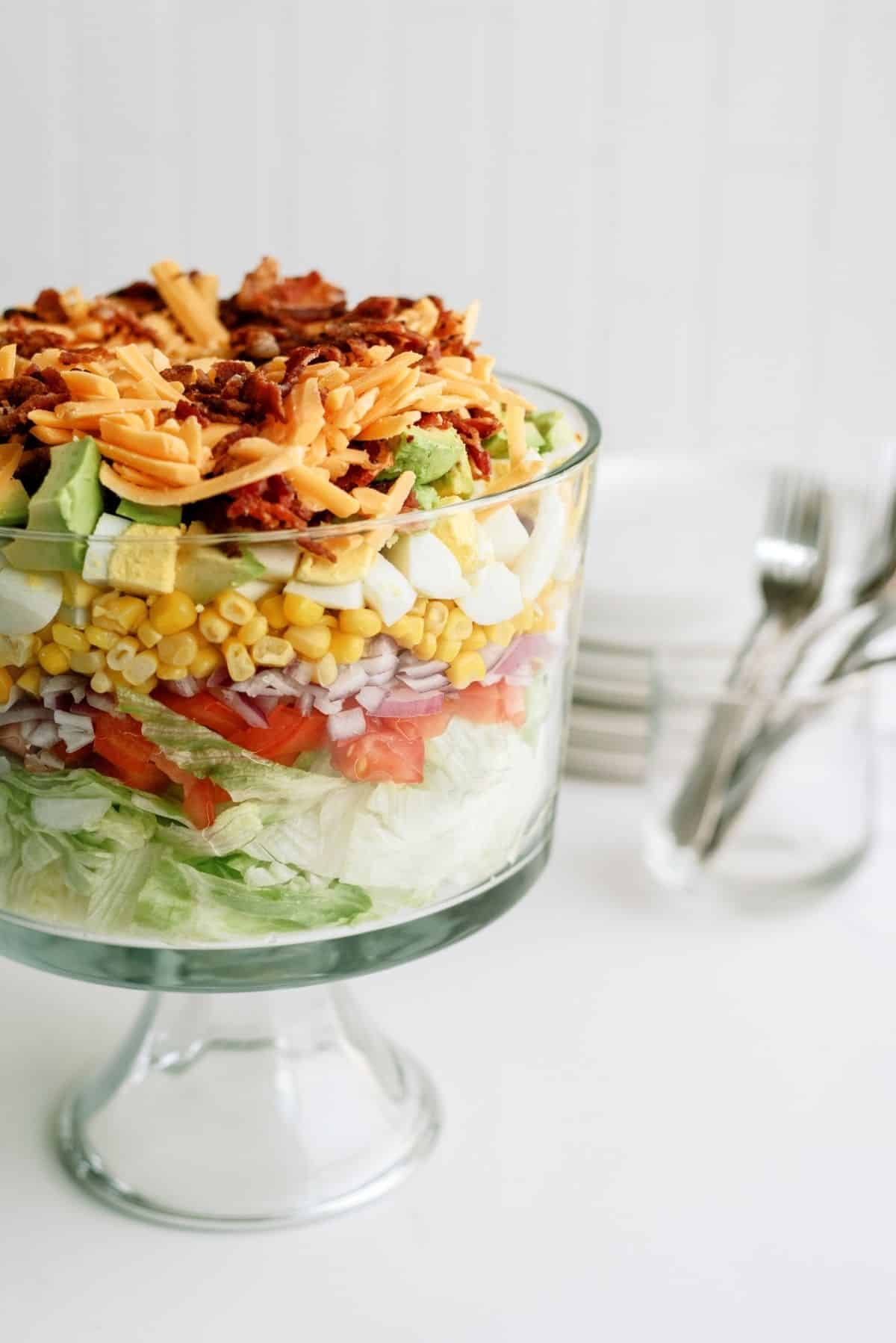Layered salad in a clear glass trifle bowl with lettuce, tomato, onion, corn, eggs, avocado, shredded cheddar cheese, and bacon. Plates and forks are in the background.