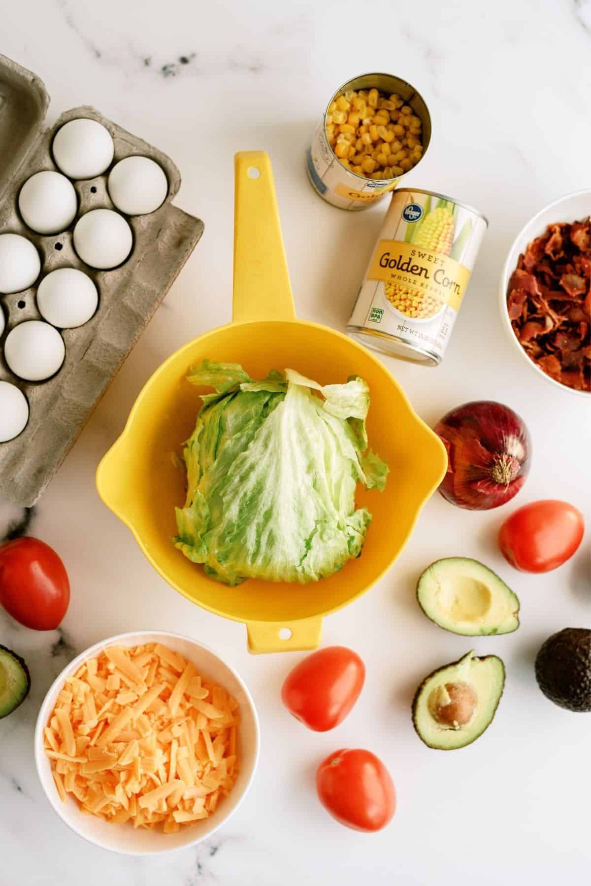Ingredients for a salad are arranged on a white surface, including eggs, lettuce, canned corn, bacon, red onion, tomatoes, avocado, and shredded cheddar cheese.