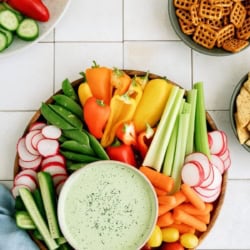 A platter of assorted fresh vegetables with a bowl of green dip in the center, surrounded by bowls of pretzels, chips, and sliced cucumbers on a white tiled surface.