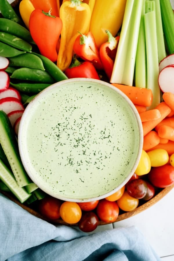 A bowl of green dipping sauce garnished with herbs, surrounded by assorted fresh vegetables including celery, carrots, tomatoes, radishes, cucumbers, and peppers.