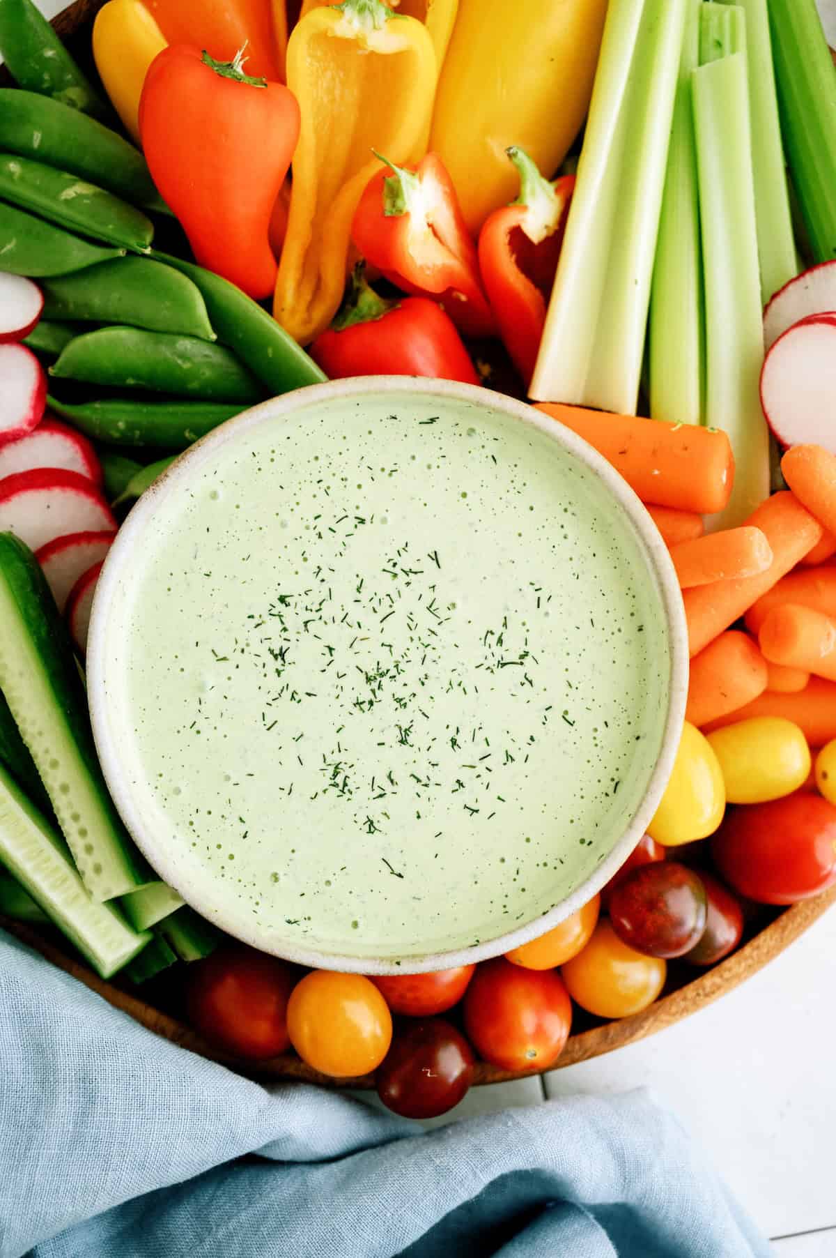A bowl of green dipping sauce garnished with herbs, surrounded by assorted fresh vegetables including celery, carrots, tomatoes, radishes, cucumbers, and peppers.