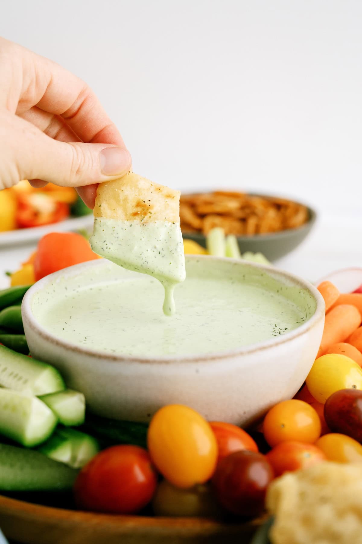 A hand dips a pita chip into a bowl of green creamy dip, surrounded by assorted fresh vegetables and tomatoes.