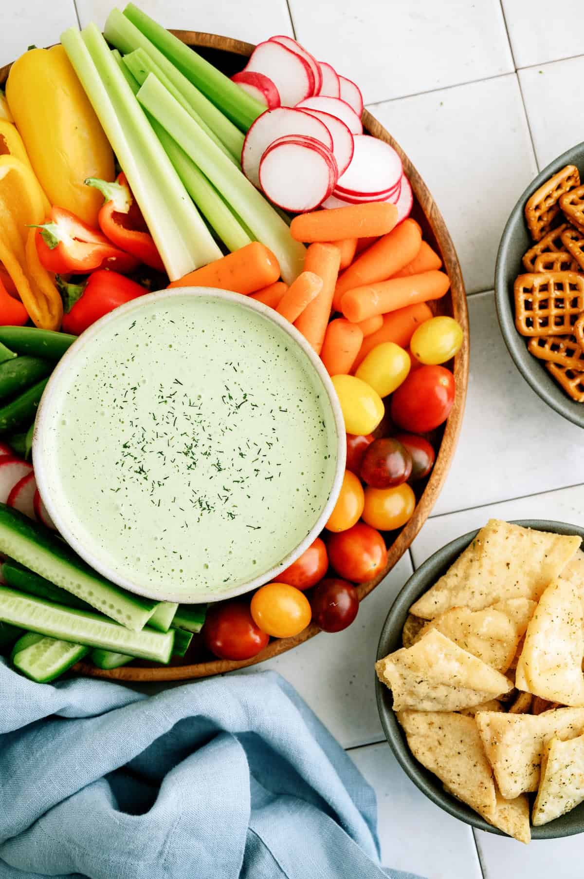 A platter of fresh vegetables, including celery, radishes, carrots, bell peppers, cucumbers, and cherry tomatoes, surrounds a bowl of green dip. Bowls of pretzels and chips are nearby.