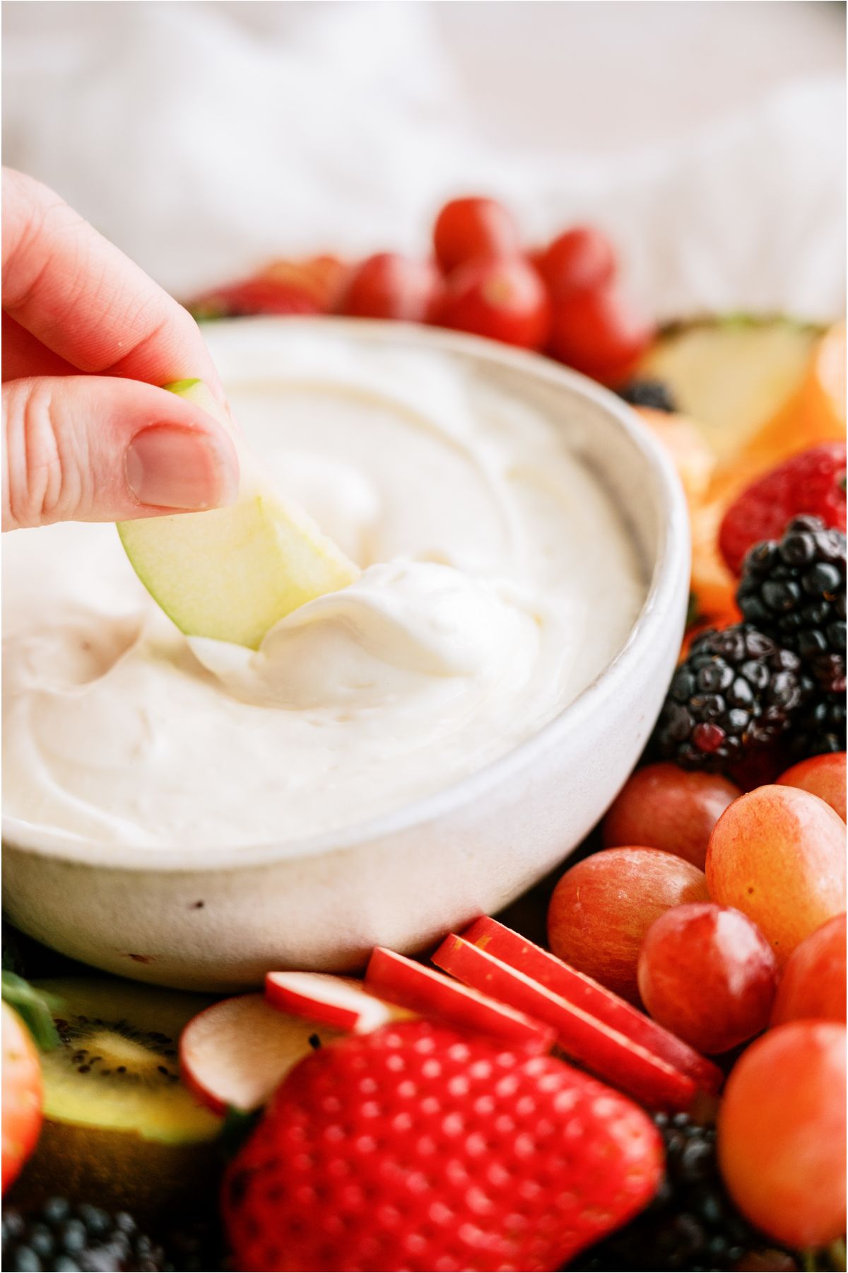 A hand dipping a sliced apple in a small bowl of Easy Cream Cheese Fruit Dip on a platter surrounded by sliced fruit.