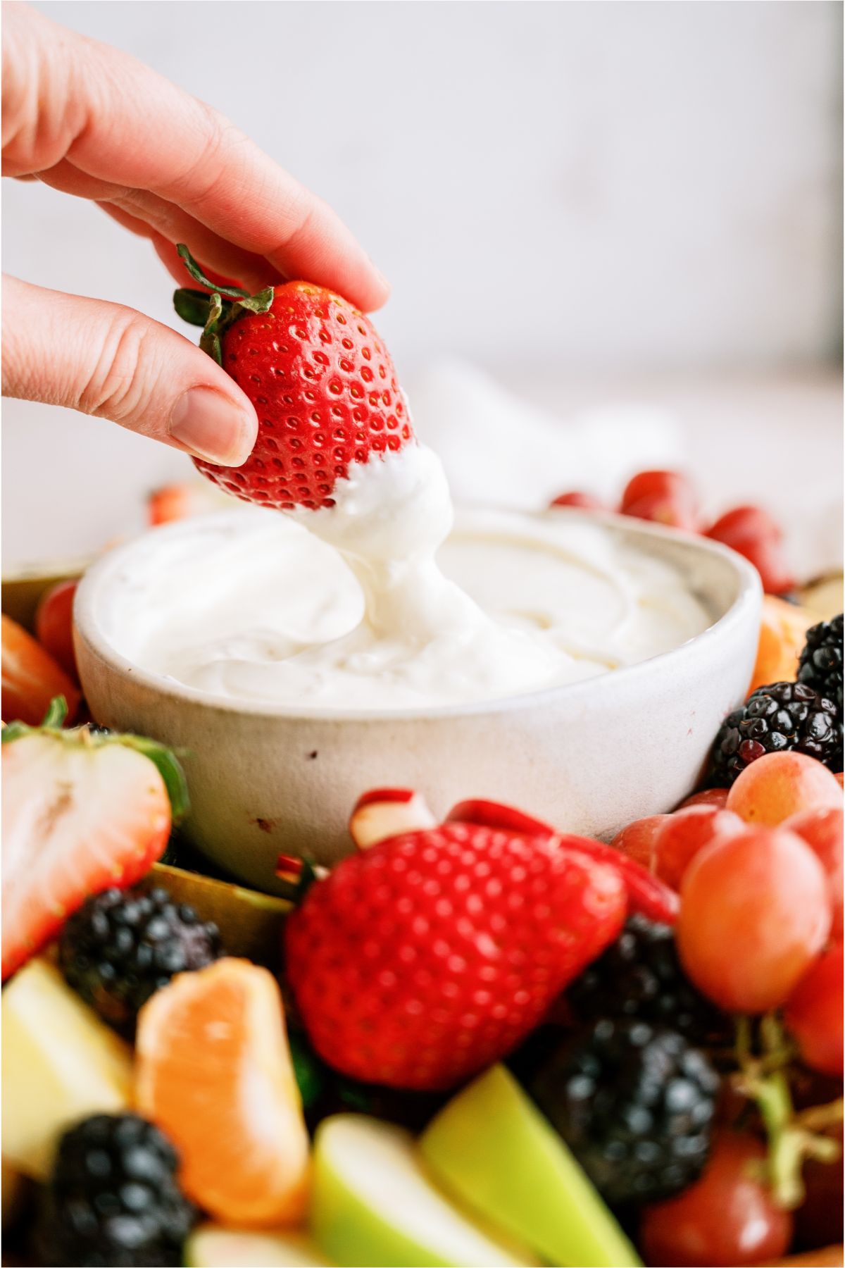 A hand dipping a fresh strawberry into a small bowl of Easy Cream Cheese Fruit Dip on a fruit platter surrounded by sliced fruit.