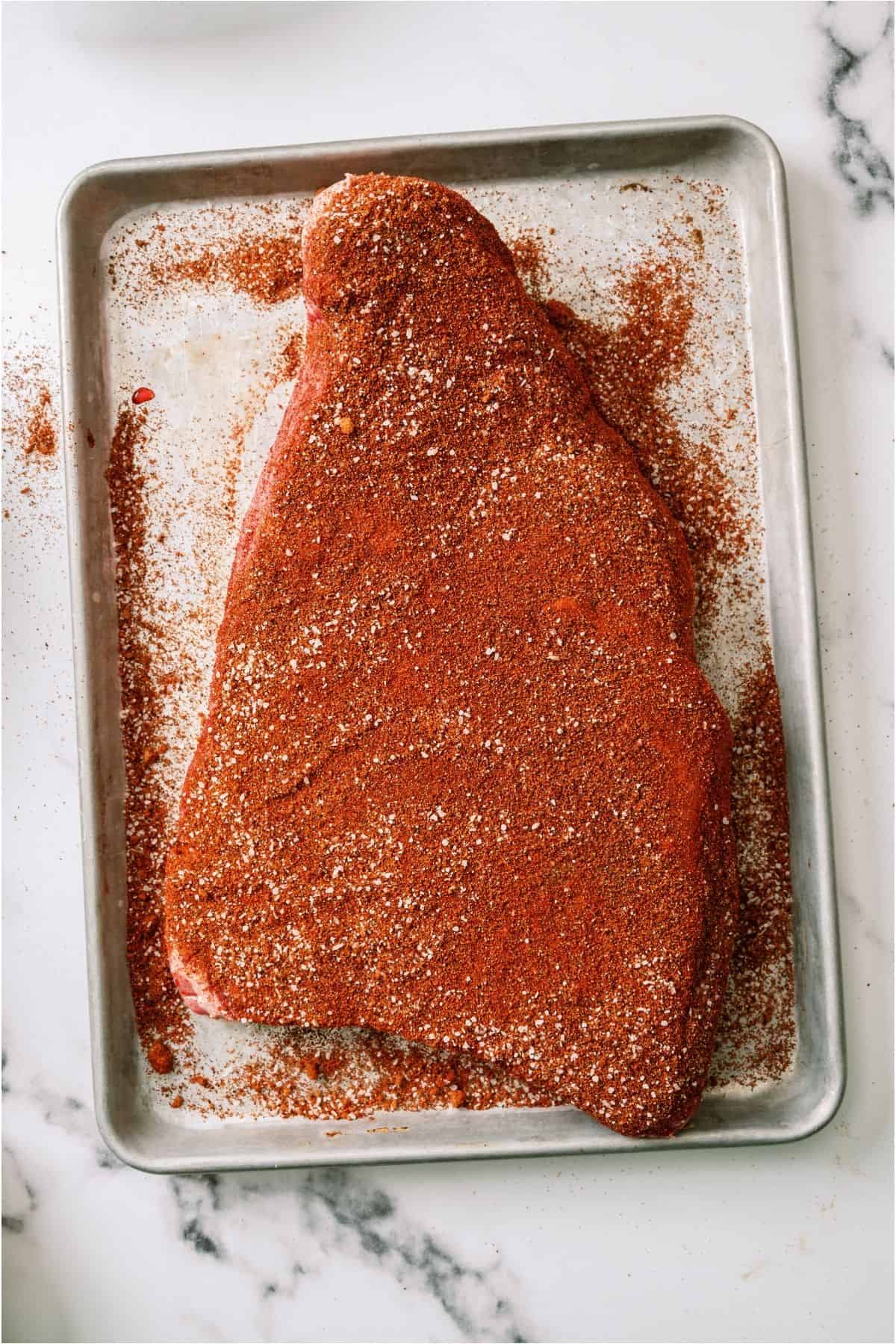 Raw Brisket covered in rub on a sheet pan.