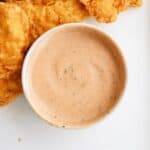 A bowl of creamy dipping sauce is placed next to several pieces of crispy, golden fried chicken strips on a white surface.