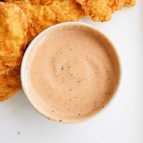 A bowl of creamy dipping sauce is placed next to several pieces of crispy, golden fried chicken strips on a white surface.