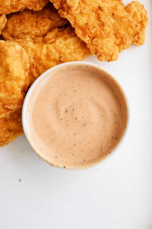A bowl of creamy dipping sauce is placed next to several pieces of crispy, golden fried chicken strips on a white surface.