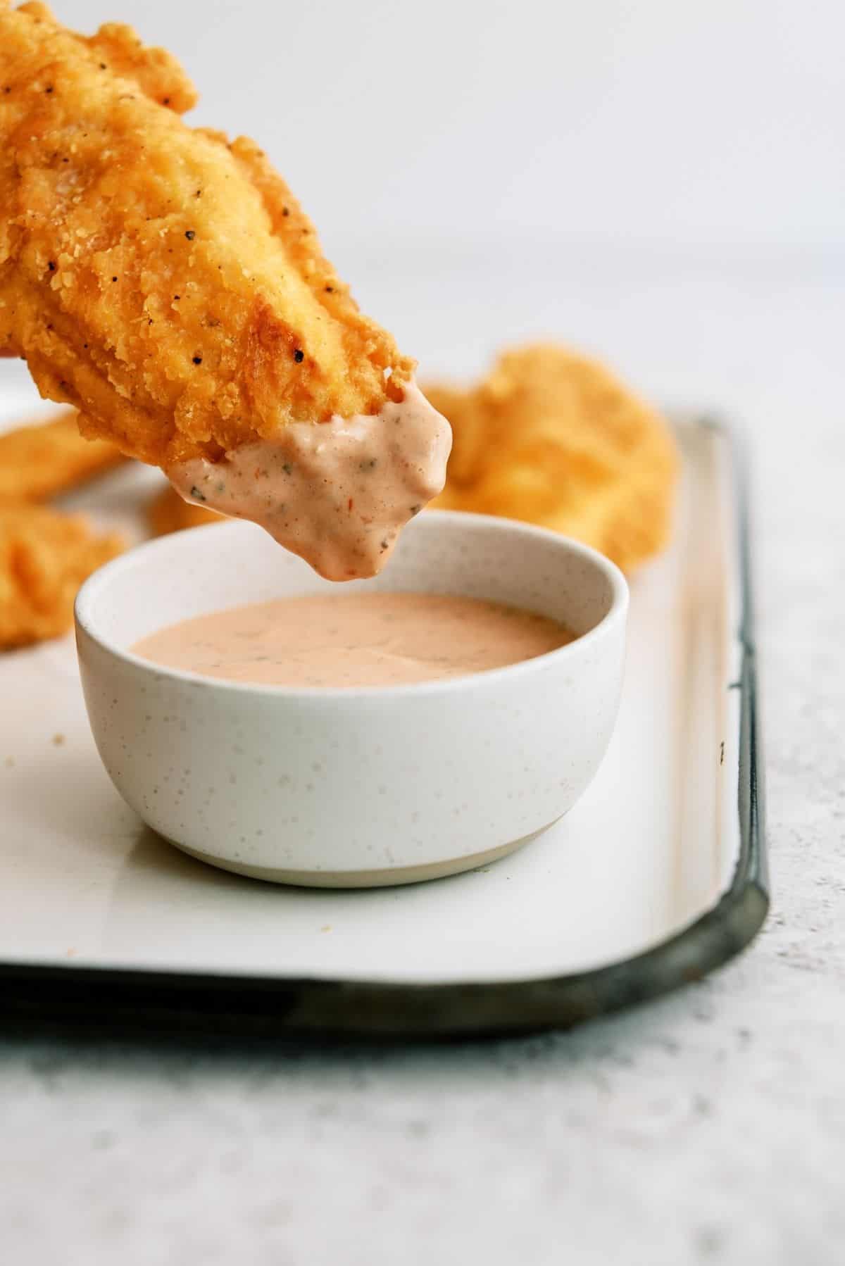 A chicken tender being dipped in a bowl of Raising Cane’s Chicken Sauce