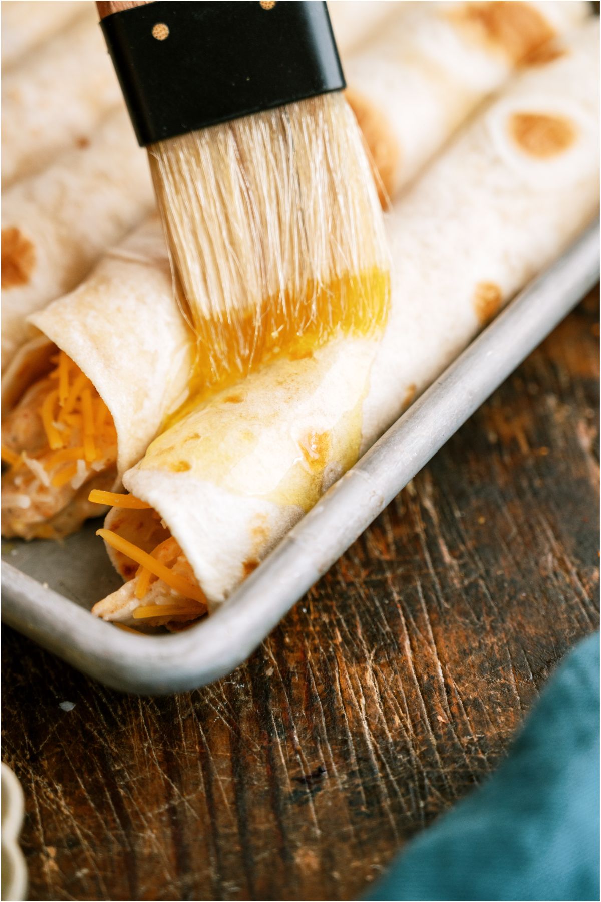 Brushing the top of unbaked Slow Cooker Cheesy Chicken Taquitos with butter on a baking sheet.