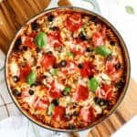 A baked pasta dish in a round pan topped with melted cheese, pepperoni slices, black olives, and fresh basil leaves on a wooden board.