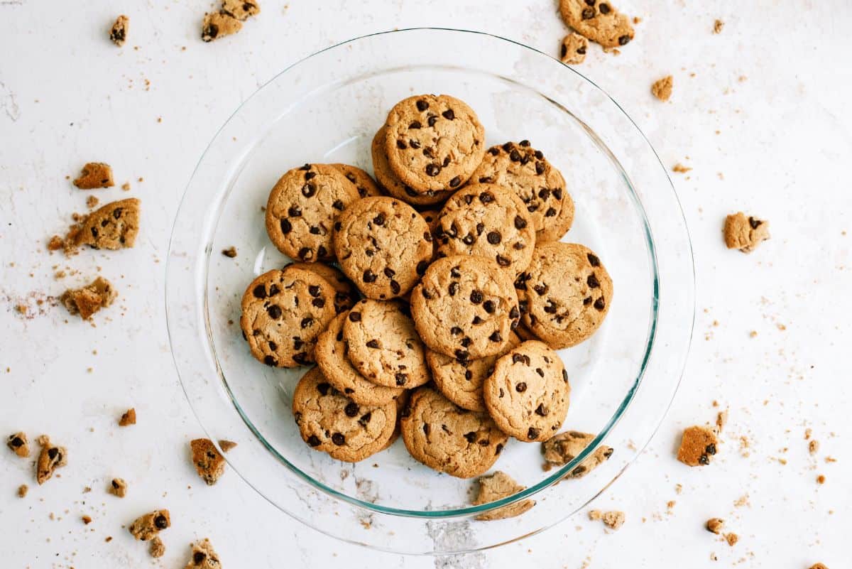 A clear glass plate with a stack of chocolate chip cookies, surrounded by scattered cookie crumbs on a white surface.