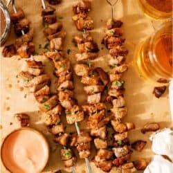 Four grilled meat skewers on parchment paper, garnished with chopped herbs, served with dipping sauce, parsley, whole garlic bulbs, and two glasses of beer.