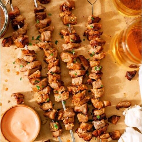 Four grilled meat skewers on parchment paper, garnished with chopped herbs, served with dipping sauce, parsley, whole garlic bulbs, and two glasses of beer.