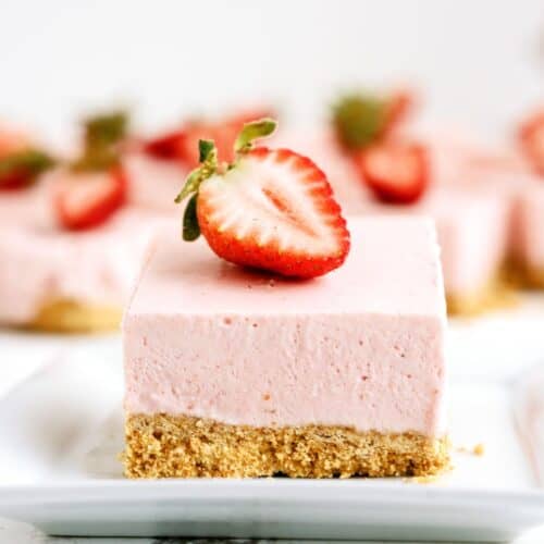 A No Bake Strawberry Cheesecake Bar on a plate with a slice of strawberry on top. More Cheesecake bars on plates in the background.